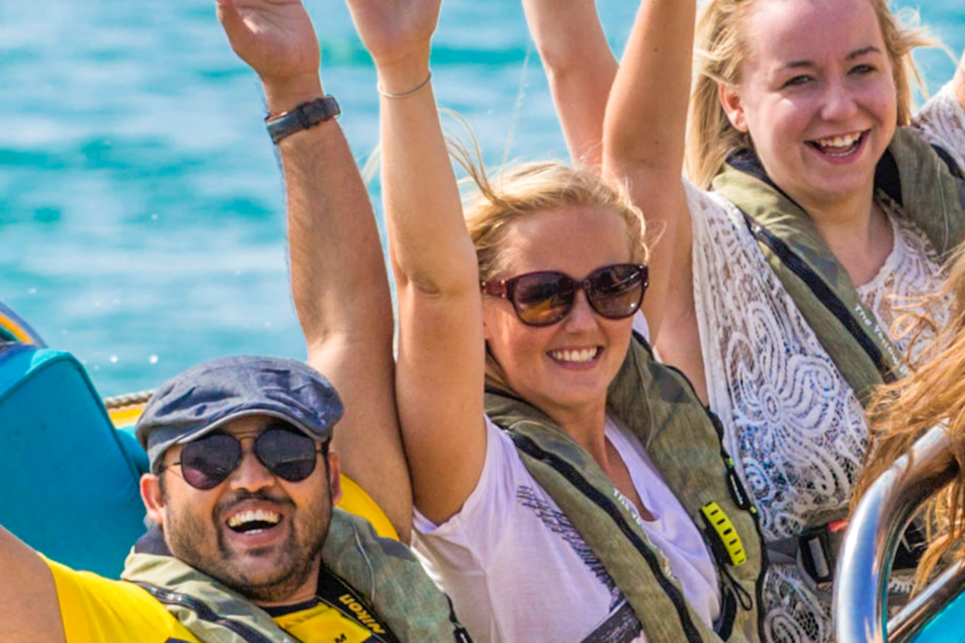 A group of people in life jackets ride a yellow speedboat with their arms raised, smiling, on a clear, sunny day at sea.