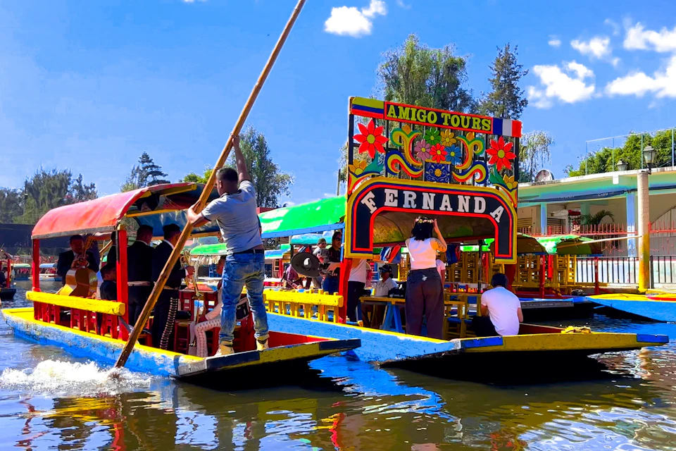 Colorful boats on a canal with people on board, one person is steering with a long pole. Sign on a boat reads "Amigo Tours Fernanda."