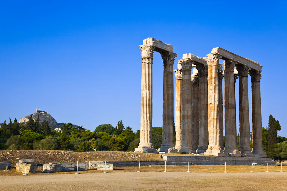 Ancient Greek temple ruins with six large columns under a clear blue sky, surrounded by trees and a fence in the foreground.
