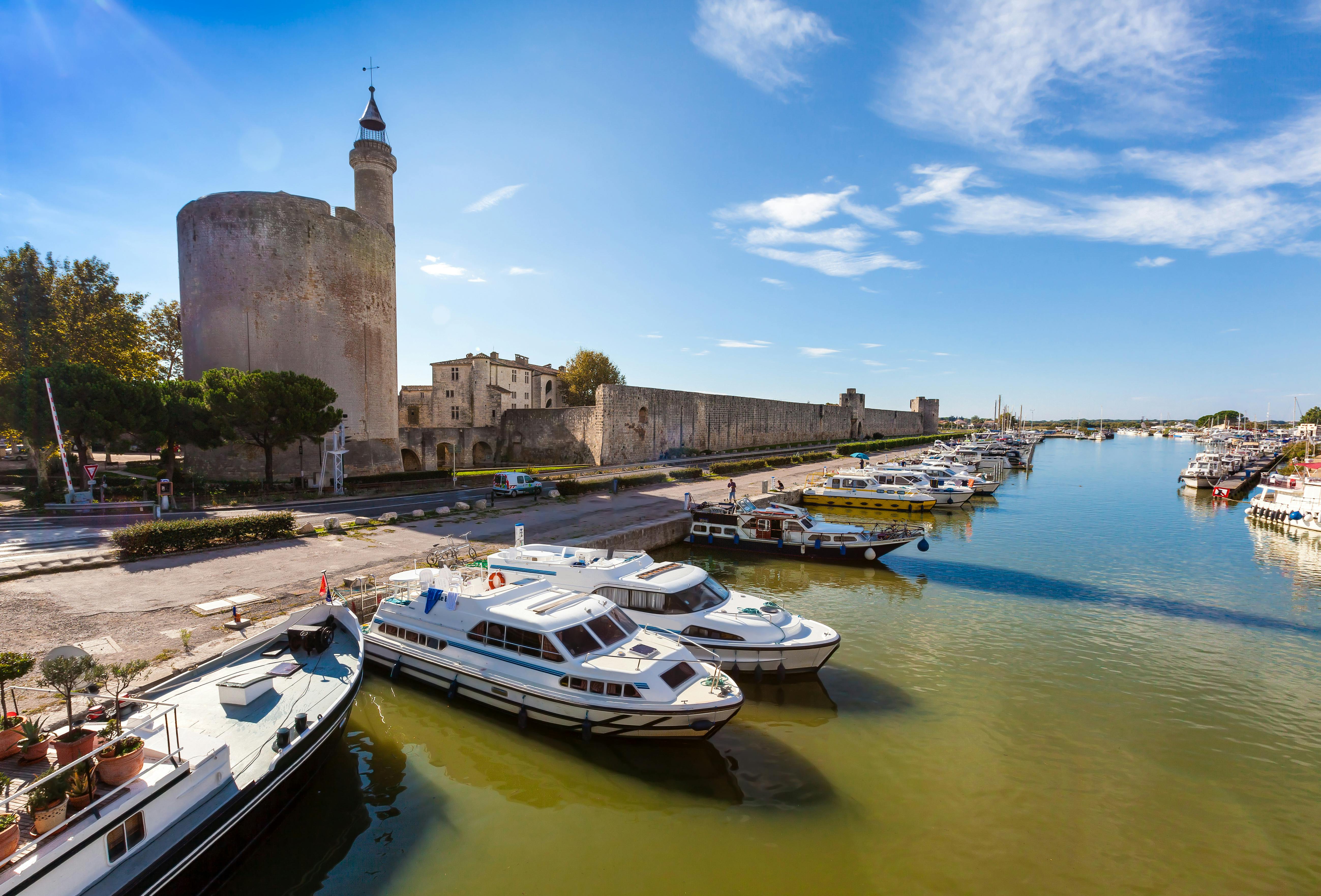 Boats docked along a waterfront with a stone castle and fortress wall in the background under a clear blue sky.