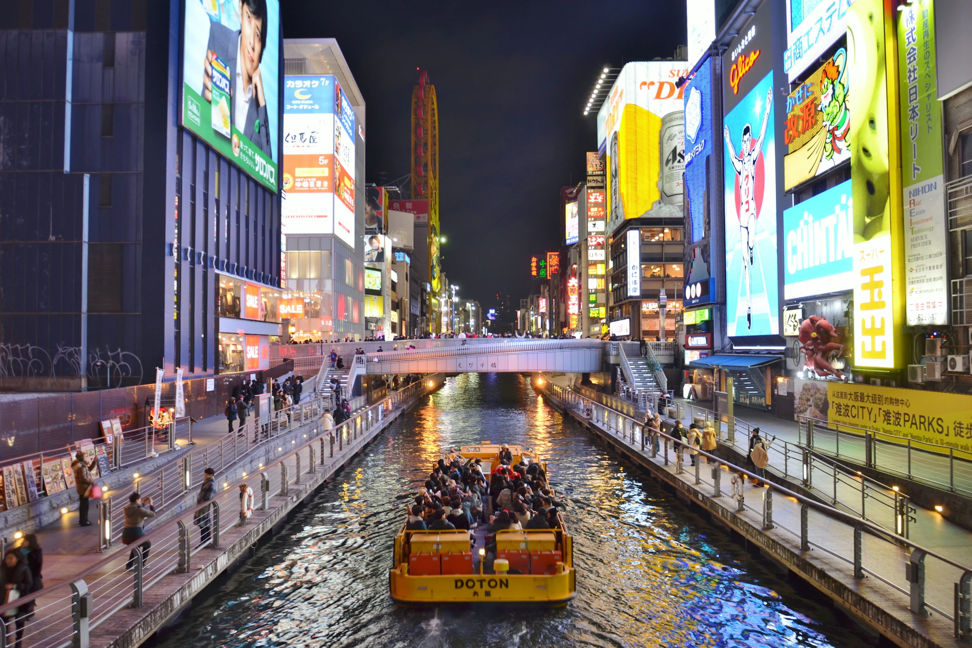 A canal in a vibrant city at night with illuminated signs, a yellow boat carrying passengers, and walkways along both sides.