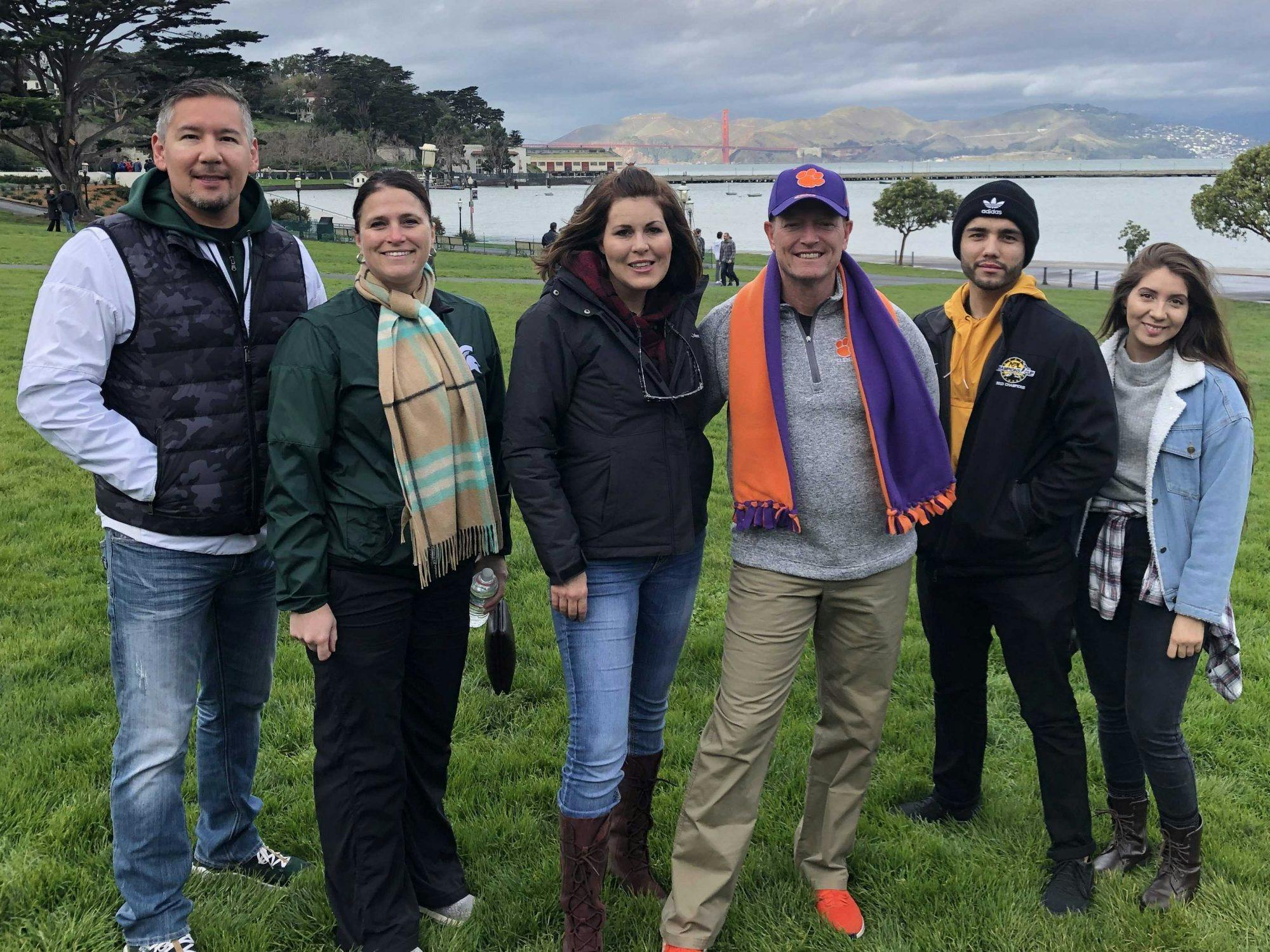 Five people standing on a grassy area near water, with the Golden Gate Bridge and hills visible in the background.