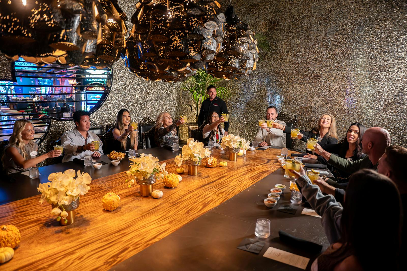 En gruppe mennesker, der sidder omkring et træbord i luksuriøse omgivelser og hæver briller i en skål med en dekorativ lysekrone over.