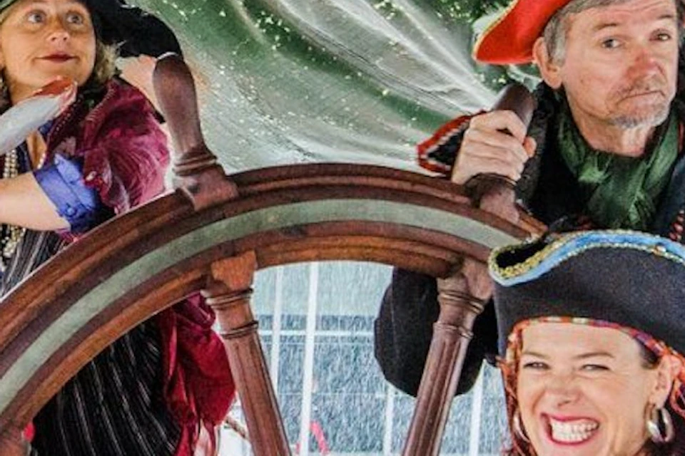 Three people dressed as pirates are gathered around a ship's wheel, with a green sail in the background.