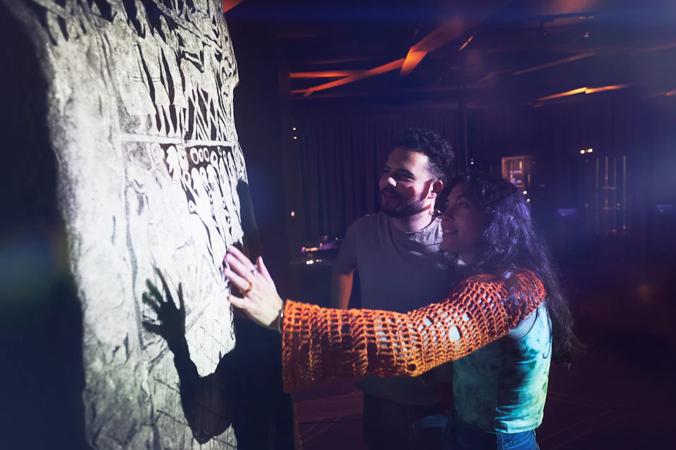 Two people examining a carved stone wall in a dimly lit indoor setting, with one person touching the carvings.
