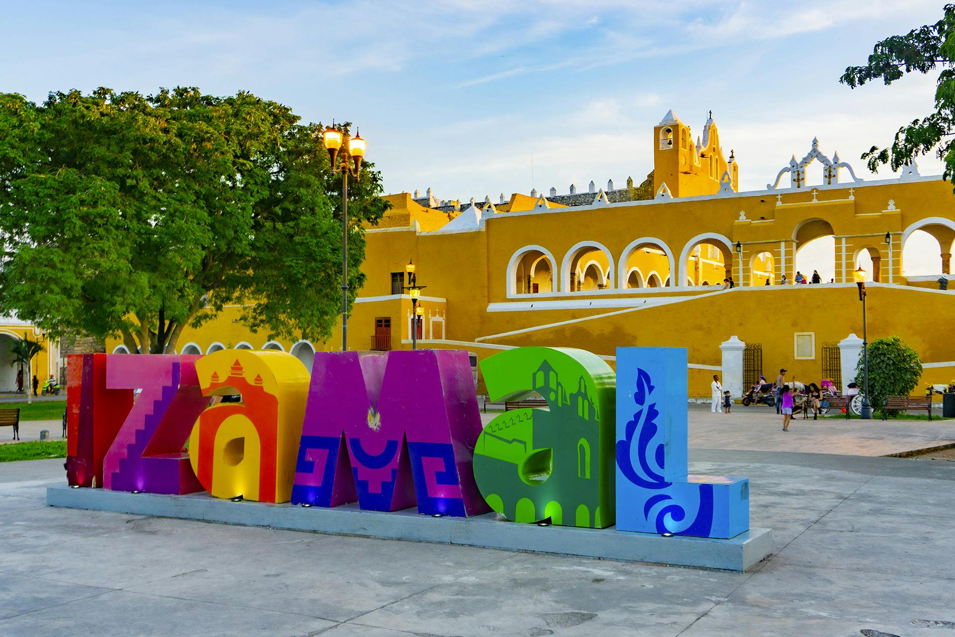 Lletres de colors "Izamal" davant d'un edifici colonial groc amb arcs, un arbre i gent en una plaça del poble.