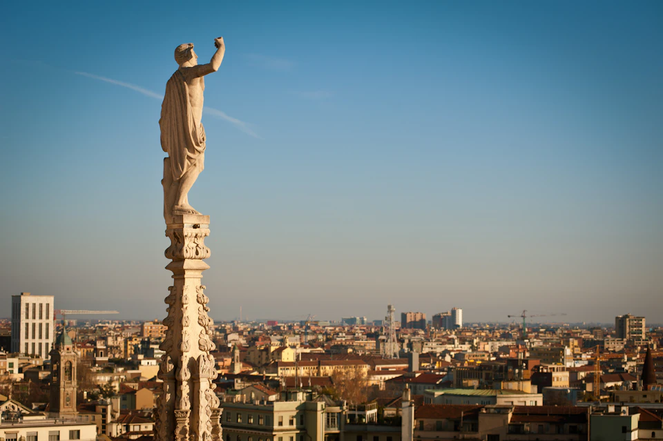 A statue of a robed figure stands atop an ornate column, overlooking a cityscape under a clear blue sky.