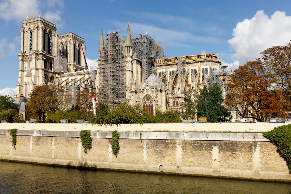 Building under reconstruction with scaffolding, seen from across a river, against a blue sky with some clouds.