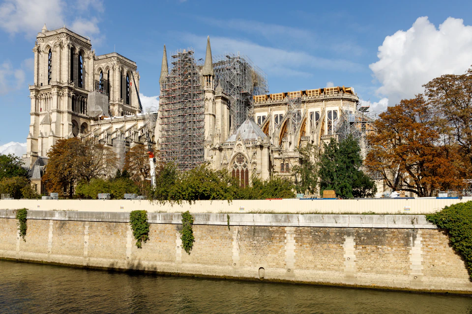 Building under reconstruction with scaffolding, seen from across a river, against a blue sky with some clouds.