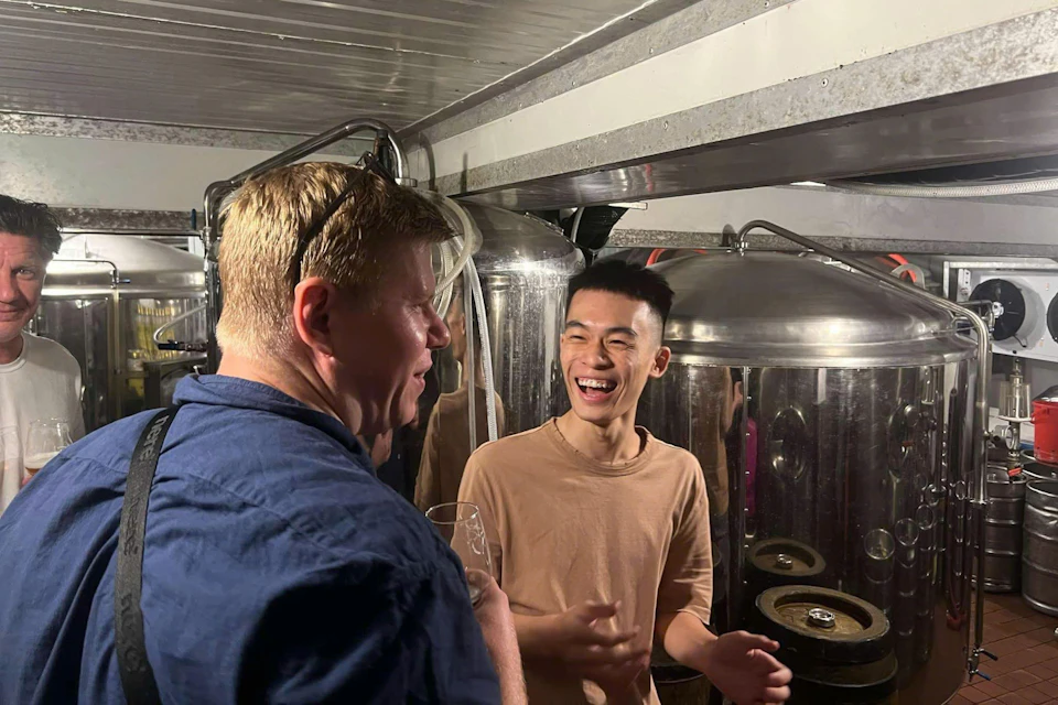 Two men laughing and talking in a brewery, surrounded by large stainless steel fermentation tanks.