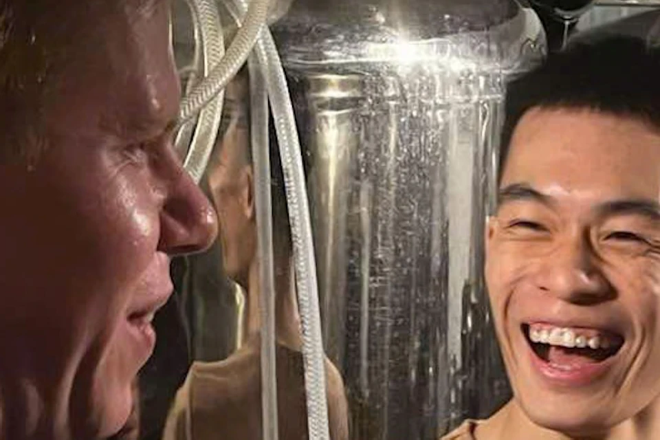 Two men laughing and talking in a brewery, surrounded by large stainless steel fermentation tanks.