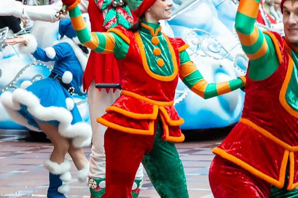 Colorful Christmas parade with people in festive costumes and roller skates, Santa in a sleigh, and onlookers watching from the side.