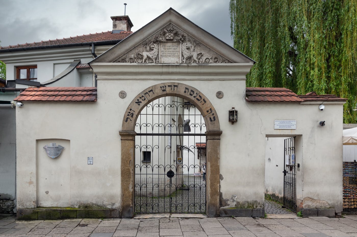 Ornate gate with Hebrew inscription, flanked by a cream-colored wall and smaller entrance, with a historic building behind.
