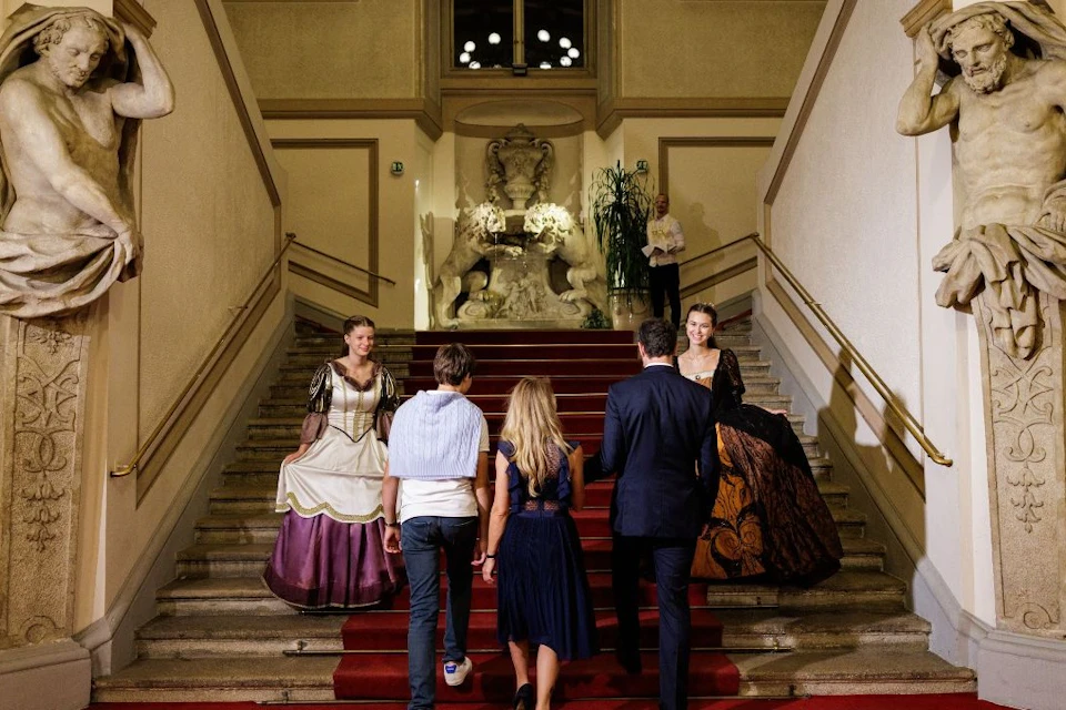 A group of people dressed in formal and historical attire ascending a red-carpeted staircase in an ornate interior setting.
