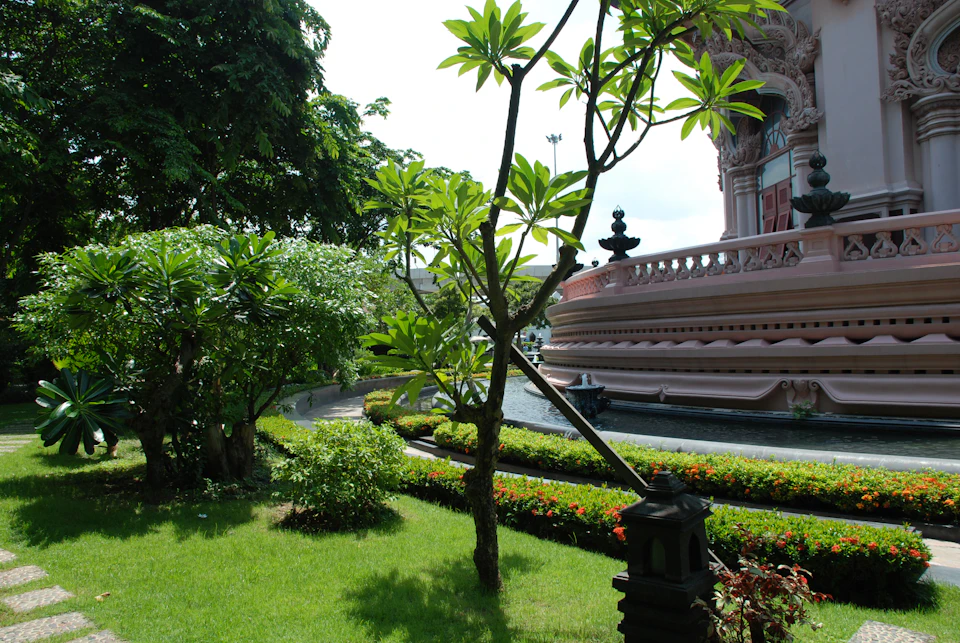 Lush garden with trees, manicured bushes, and a pathway next to an ornate, pink building with decorative elements.