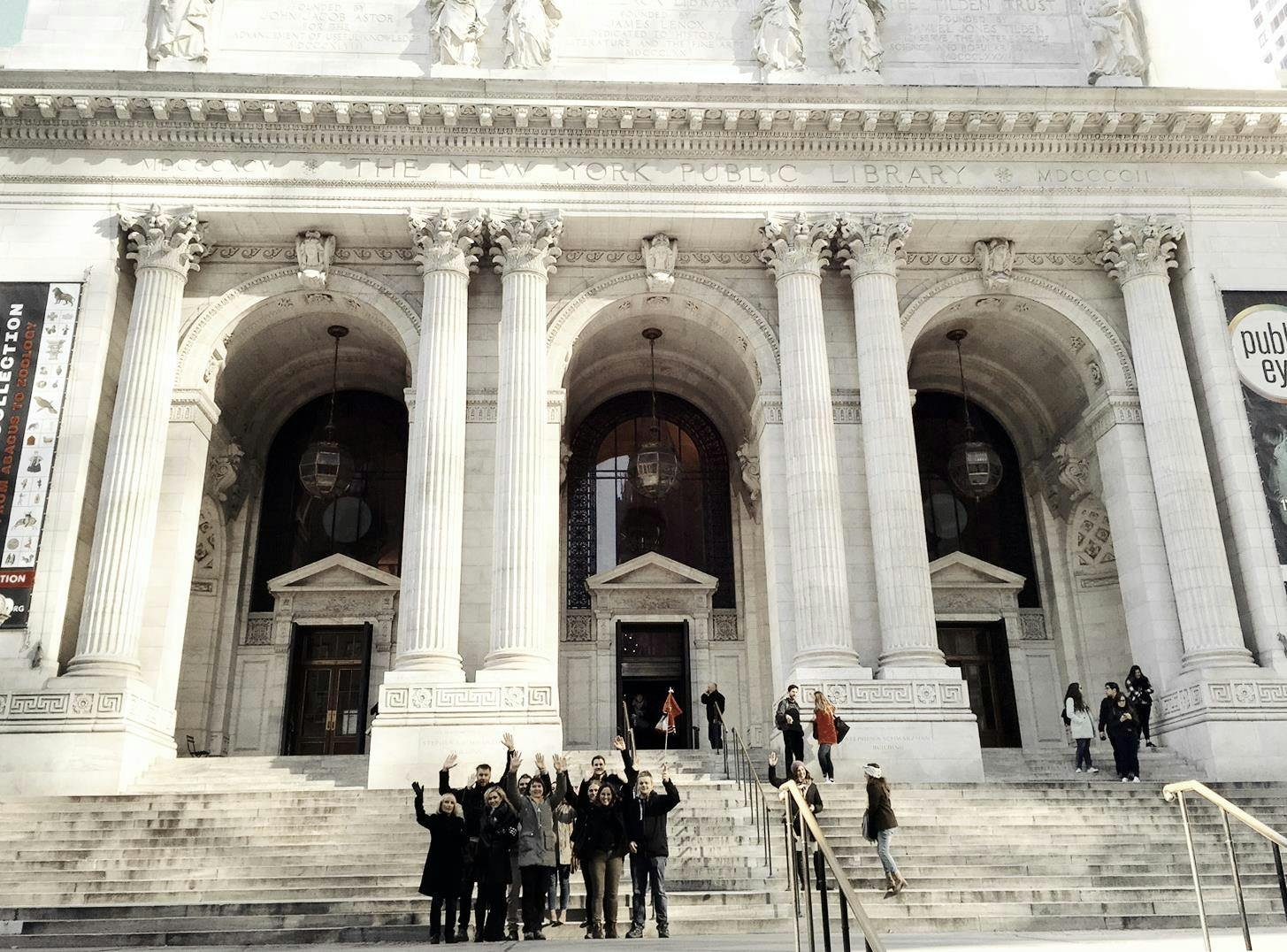 A group of people stands on the steps in front of a large building with pillars and arched entrances.
