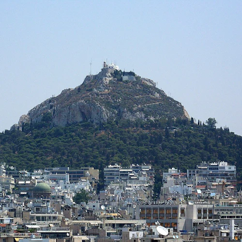 En stadsbild med många byggnader i förgrunden och en stenig kulle med strukturer på toppen i bakgrunden under en klar himmel.