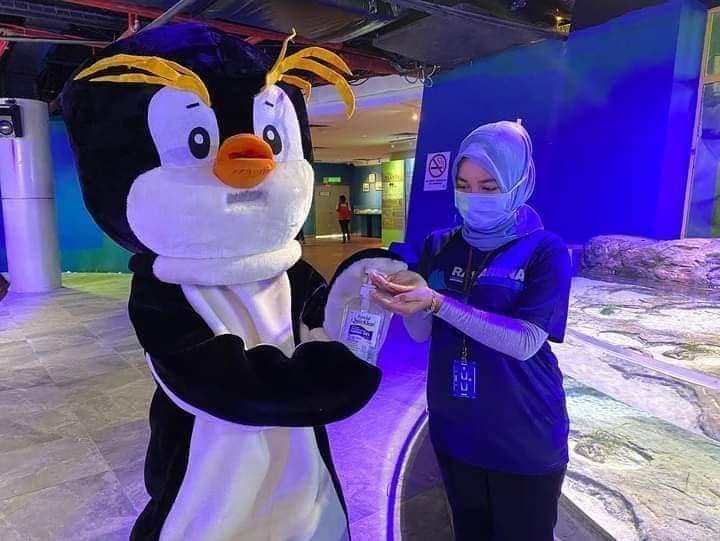 Una persona con un'uniforme blu igienizza le mani di una persona con un costume da pinguino in un ambiente chiuso e ben illuminato.