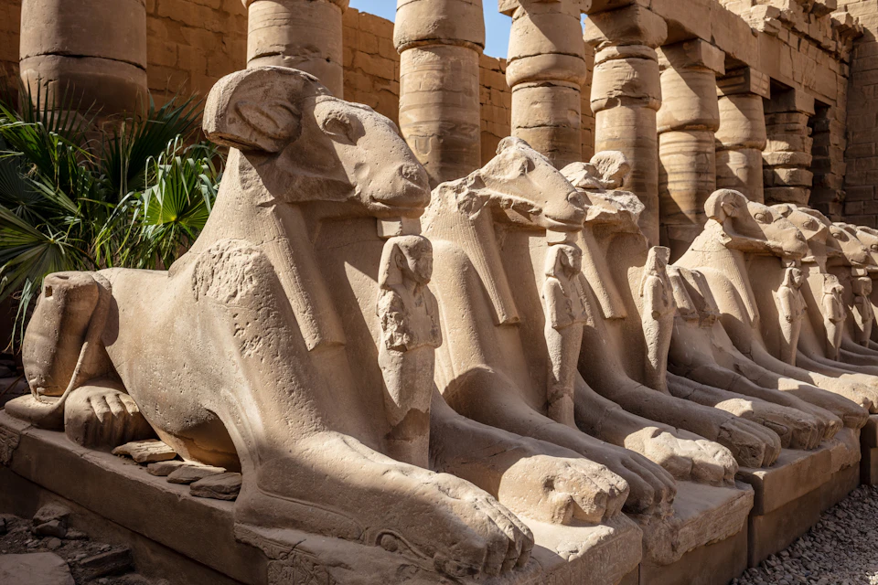 Ancient stone statues of ram-headed sphinxes in a row, with carved human figures between their legs, under a clear sky.
