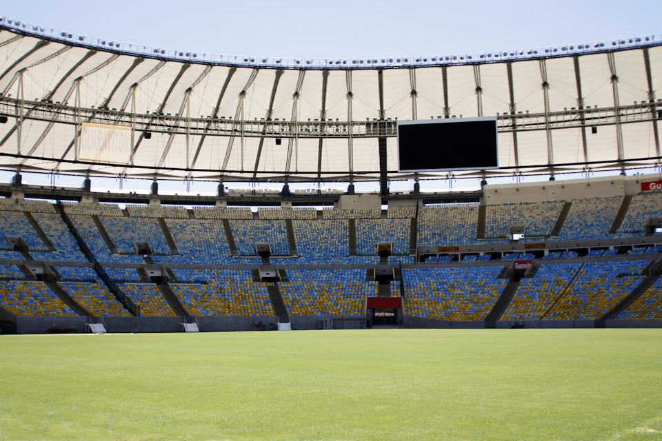 Empty stadium with blue and yellow seats, large scoreboard, and green field under a partially covered roof.
