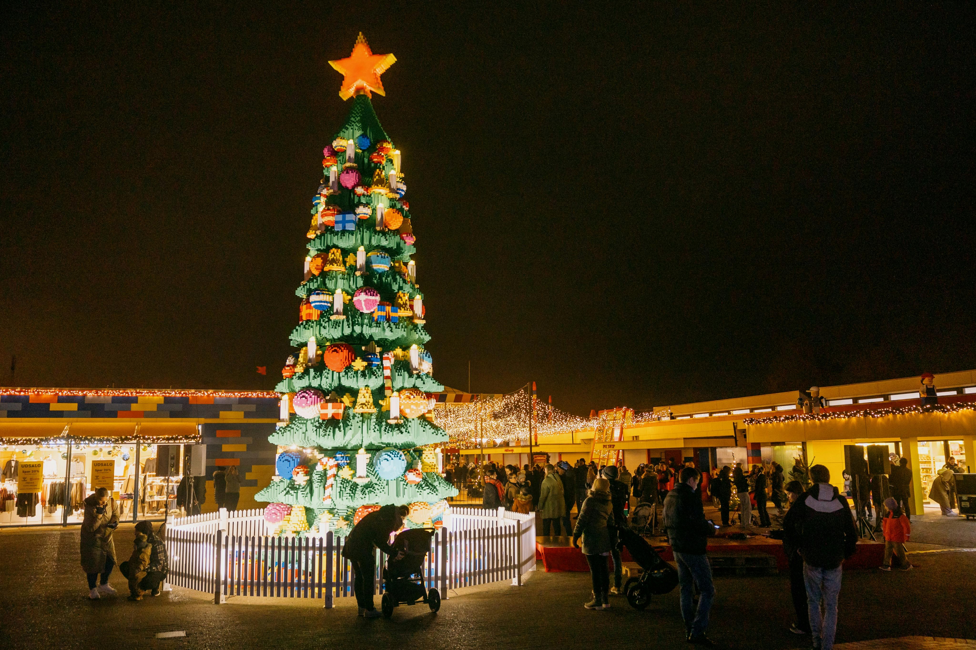 Большая, украшенная рождественская елка со звездой сверху окружена людьми и колясками, расположенными на ночном фоне.