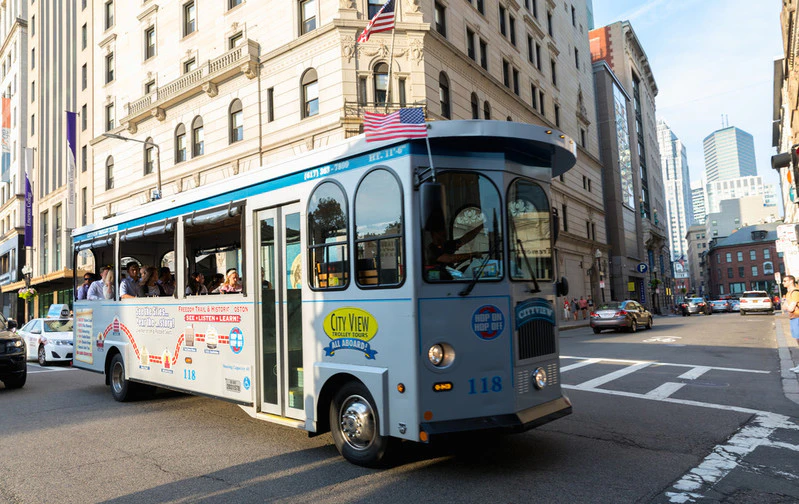 A blue and white trolley marked "City View" with passengers inside drives through an urban area with historical buildings.