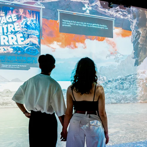 Två personer håller varandra i handen medan de tittar på en levande utställning med text och bilder relaterade till "Voyage to the Center of the Earth".
