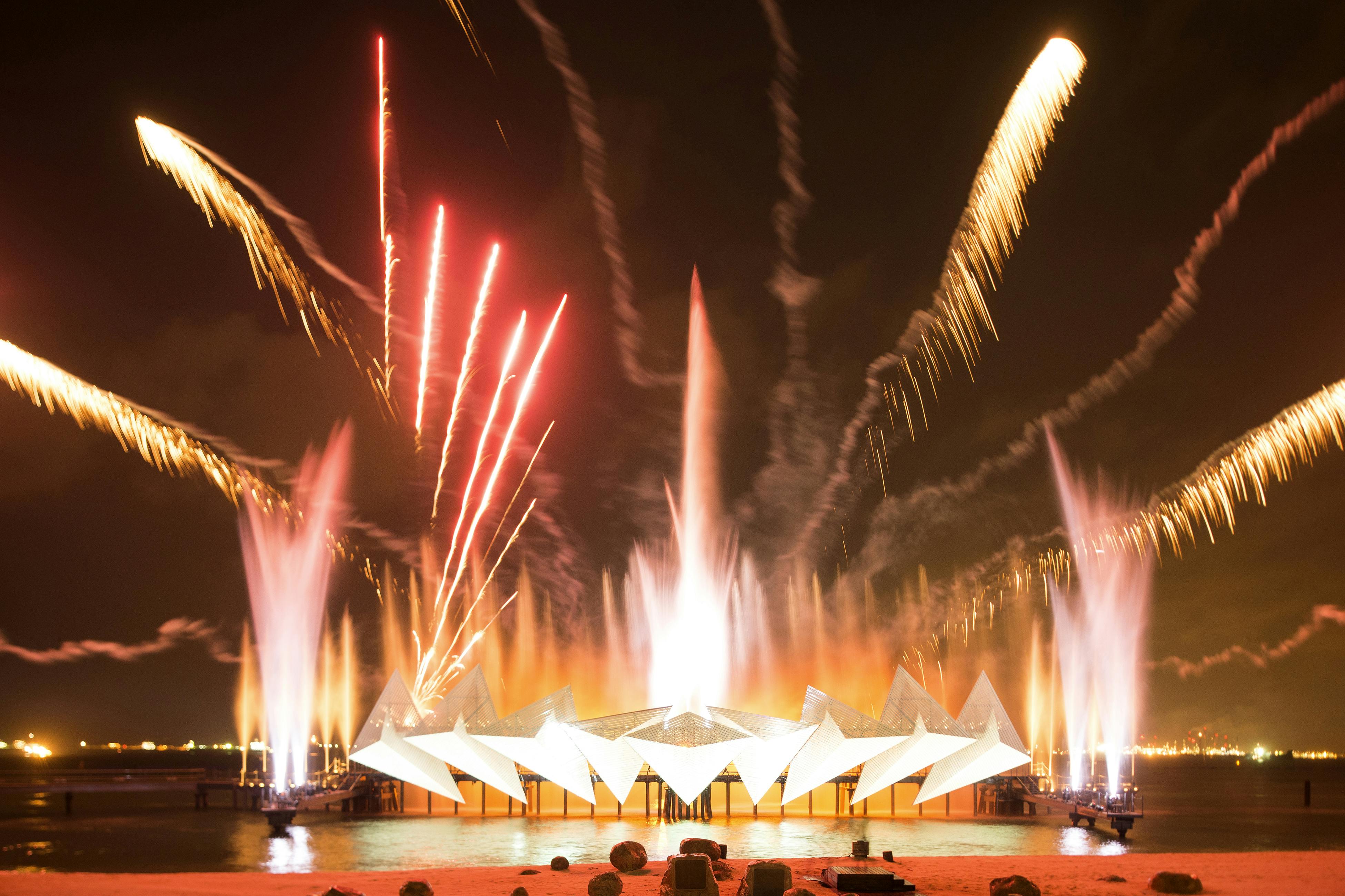 Světlý ohňostroj a barevné vodní fontány osvětlují noční oblohu kolem geometrické struktury na nábřeží.