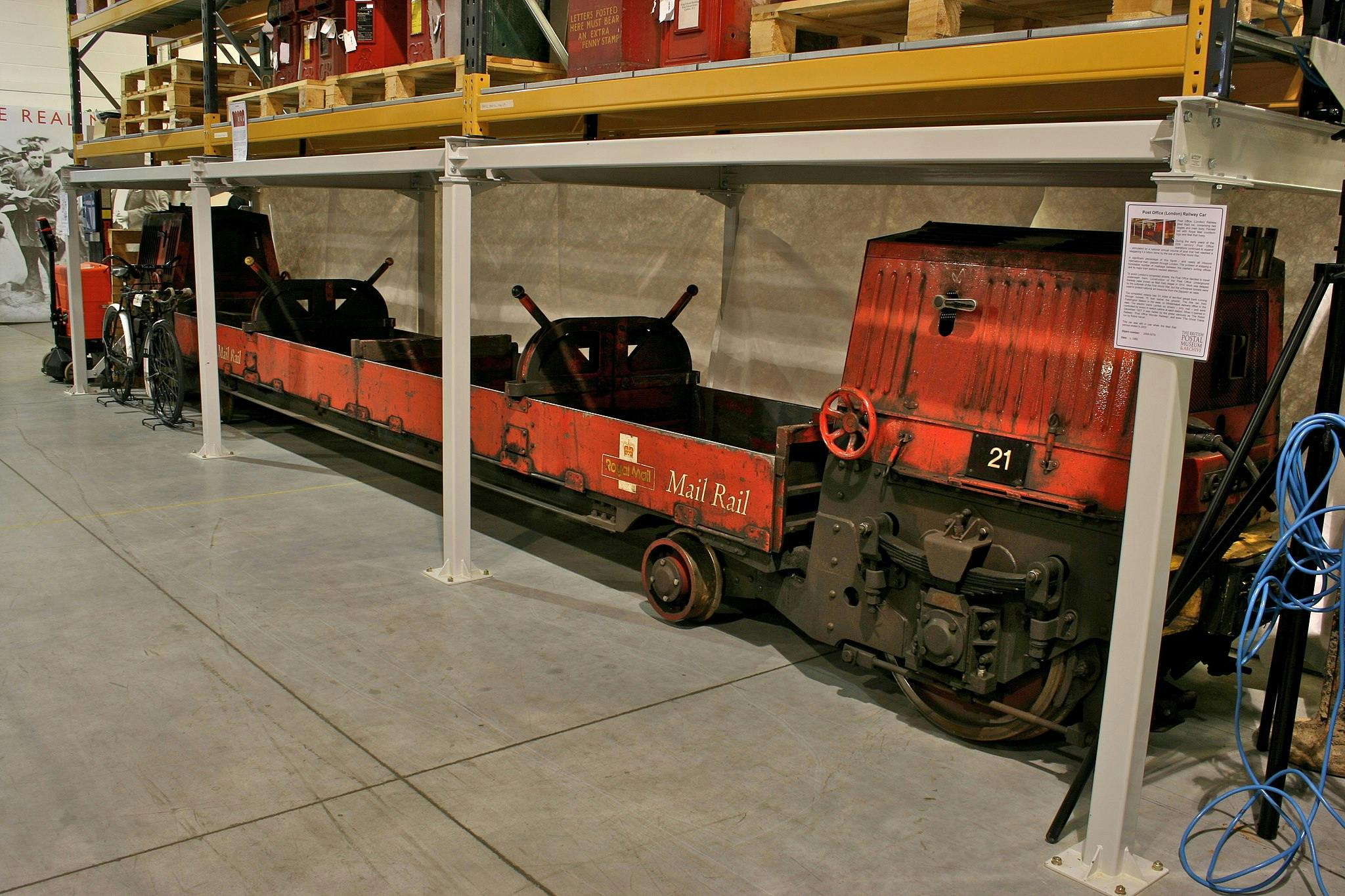 Ancien wagon rouge de Mail Rail exposé dans un entrepôt intérieur avec des étagères et d'autres objets à l'arrière-plan.