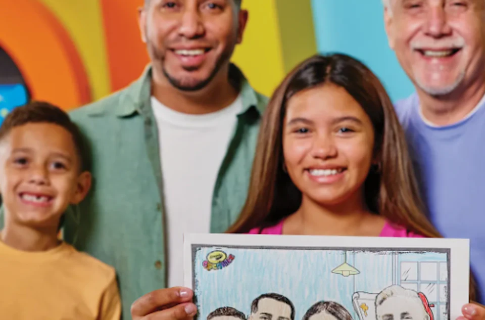 A group of four people posing, with one holding a colorful drawing of them together, standing in a brightly colored setting.