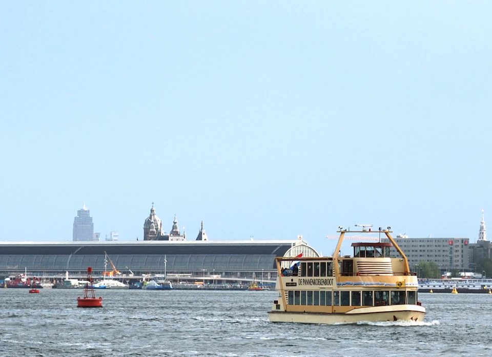 A yellow ferry named "IJ-veren Amsterdam" sails on a river with a large terminal building and city skyline in the background.