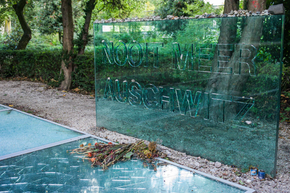 A reflective glass memorial with the inscription "Nooit Meer Auschwitz" and wilted flowers laid at its base. Trees are in the background.
