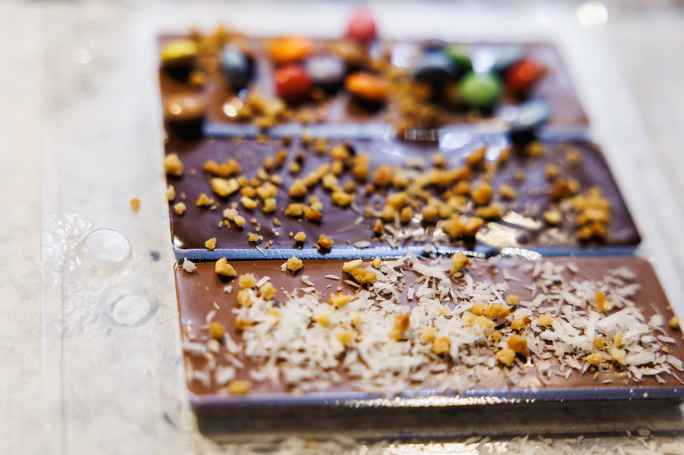 A close-up of a chocolate bar divided into sections, topped with crushed nuts and colorful candy pieces.