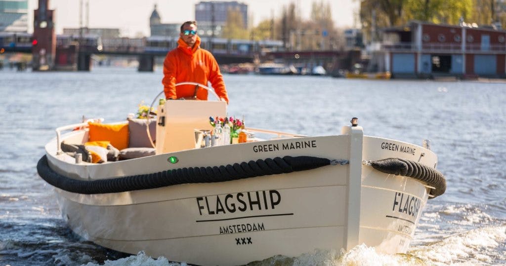 A man in an orange jacket navigates a boat labeled "Flagship Amsterdam" on a city river with a bridge and buildings in the background.