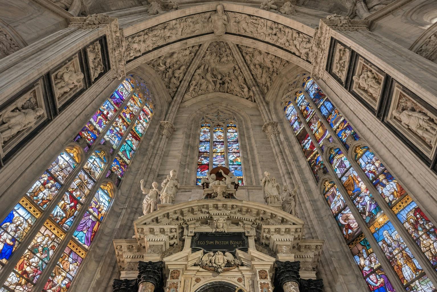 Interior of a cathedral with ornate stained glass windows, detailed carvings, and a central altar adorned with statues.