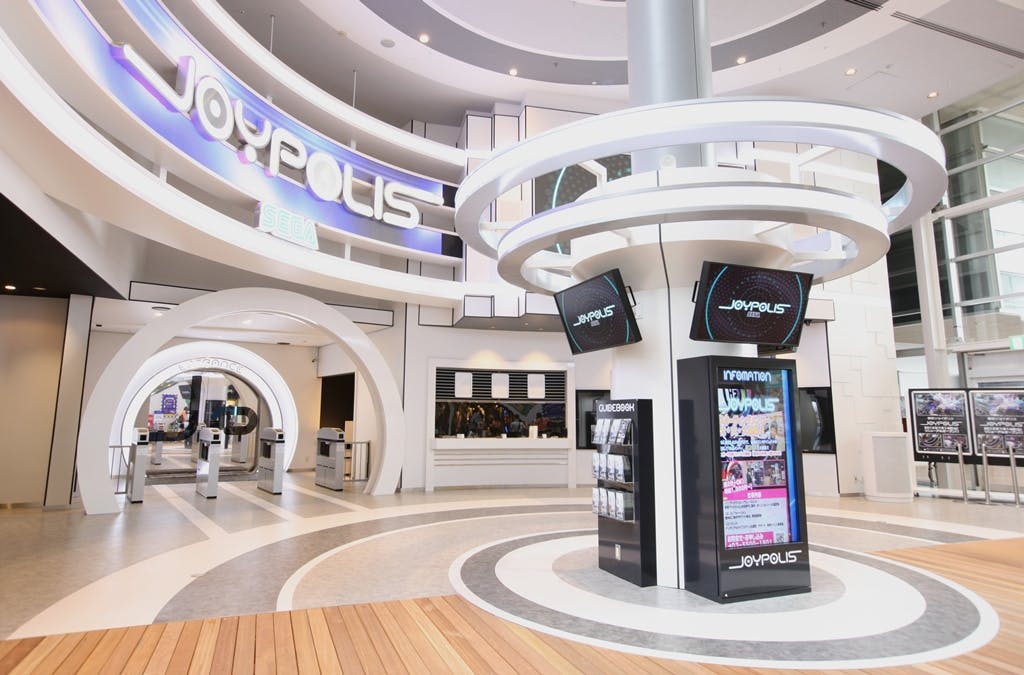 Modern indoor amusement park entrance with large "Joypolis" signage, electronic displays, ticket gates, and futuristic architecture.