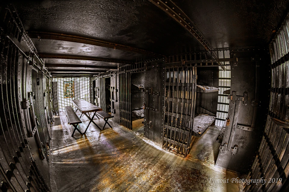 Dimly lit prison cell with metal bunk beds, a wooden table, benches, and barred doors. Floor shows wear and tear.