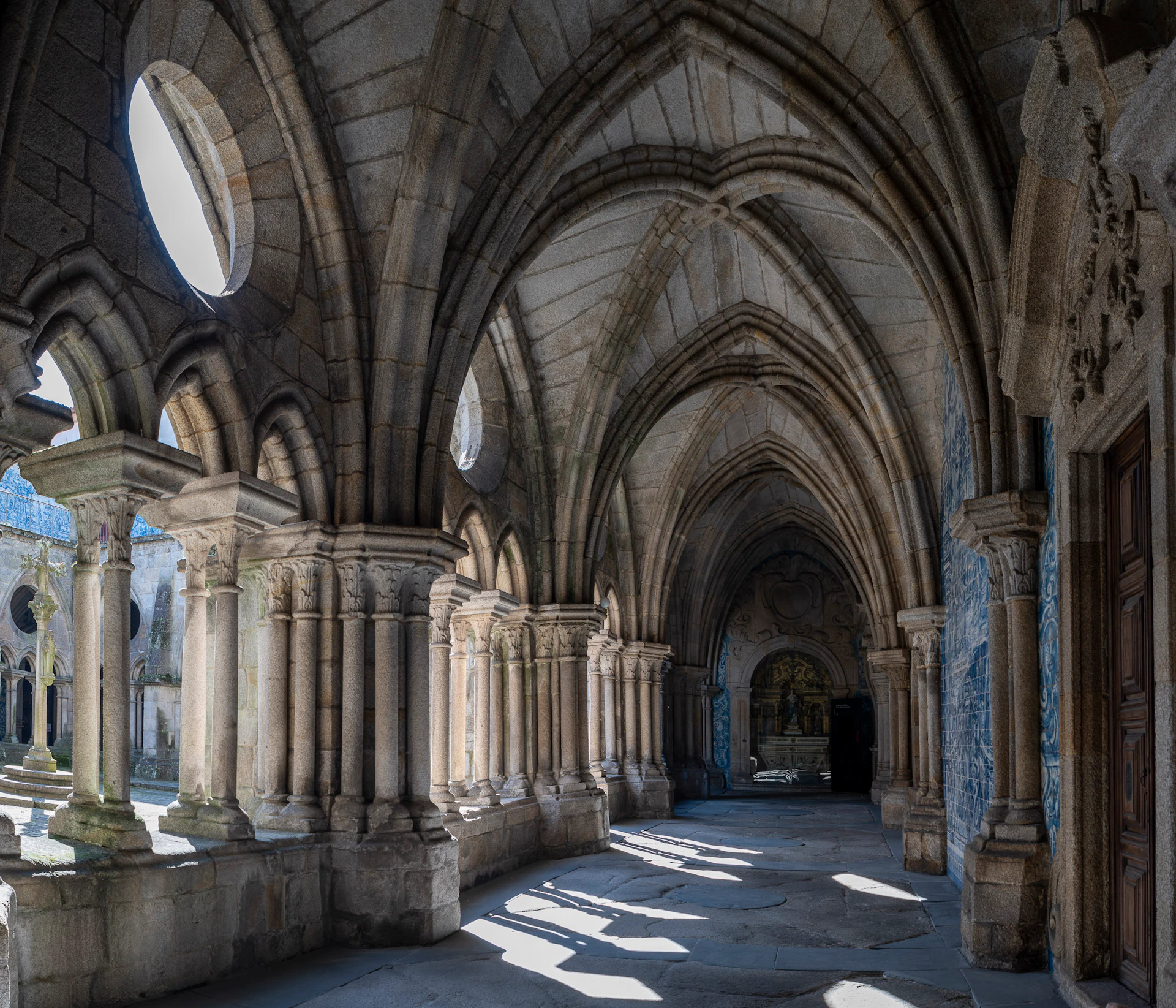 Stone cloister with arched ceiling and columns, illuminated by natural light from large windows.