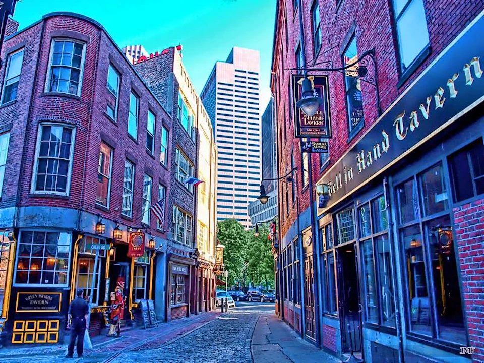 A narrow cobblestone street with historic brick buildings, a few pedestrians, and a modern skyscraper in the background.