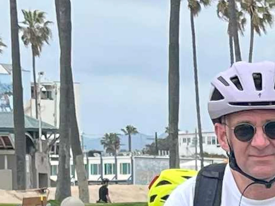 Group of cyclists posing with bicycles, wearing helmets and casual clothes, near palm trees and a beachside promenade.