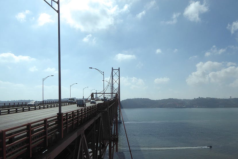 A red suspension bridge with vehicles driving across it, spanning over a body of water under a partly cloudy sky.