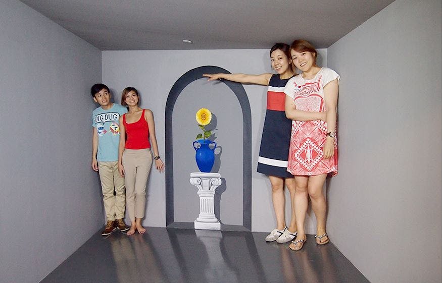 Čtyři lidé stojí v optické iluzorní místnosti s šedými stěnami a modrou vázou se žlutým květem na podstavci.