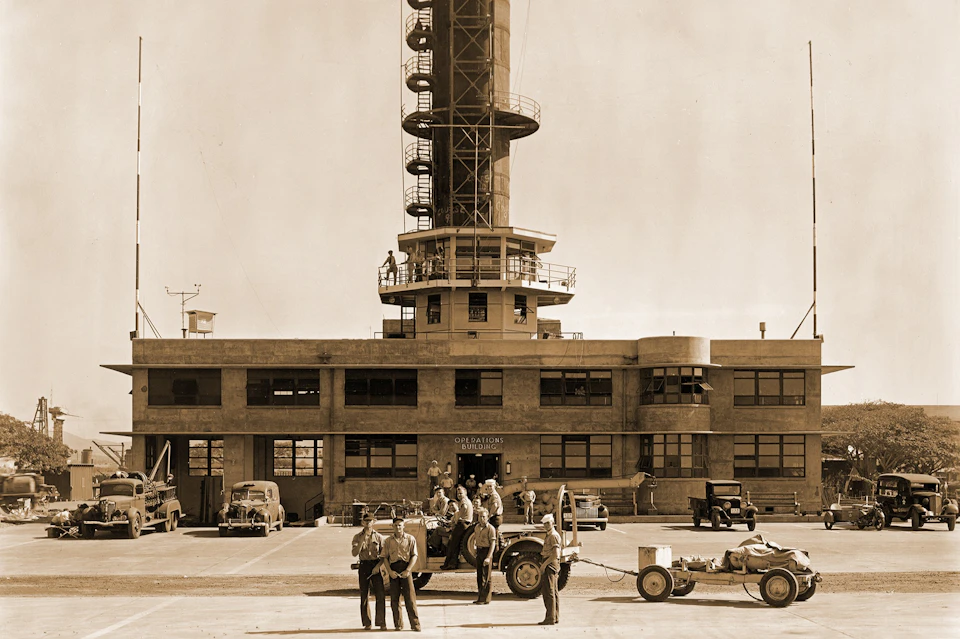 Sepia-toned image of a control tower with several military personnel and vehicles in front, including a jeep and a fuel trailer.