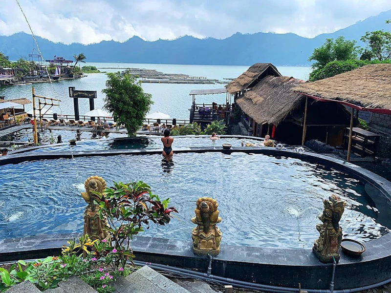 A scenic pool with stone statues and people swimming, overlooking a lake and mountains in the distance.