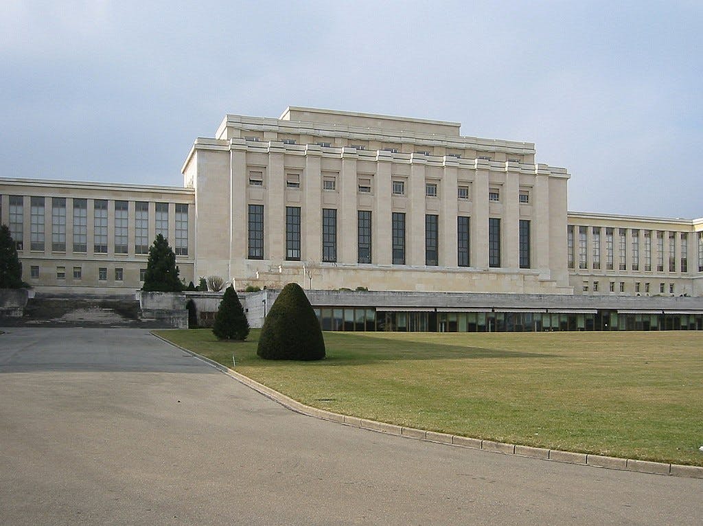A large, beige government or institutional building with tall windows, surrounded by a wide lawn and path, under a cloudy sky.