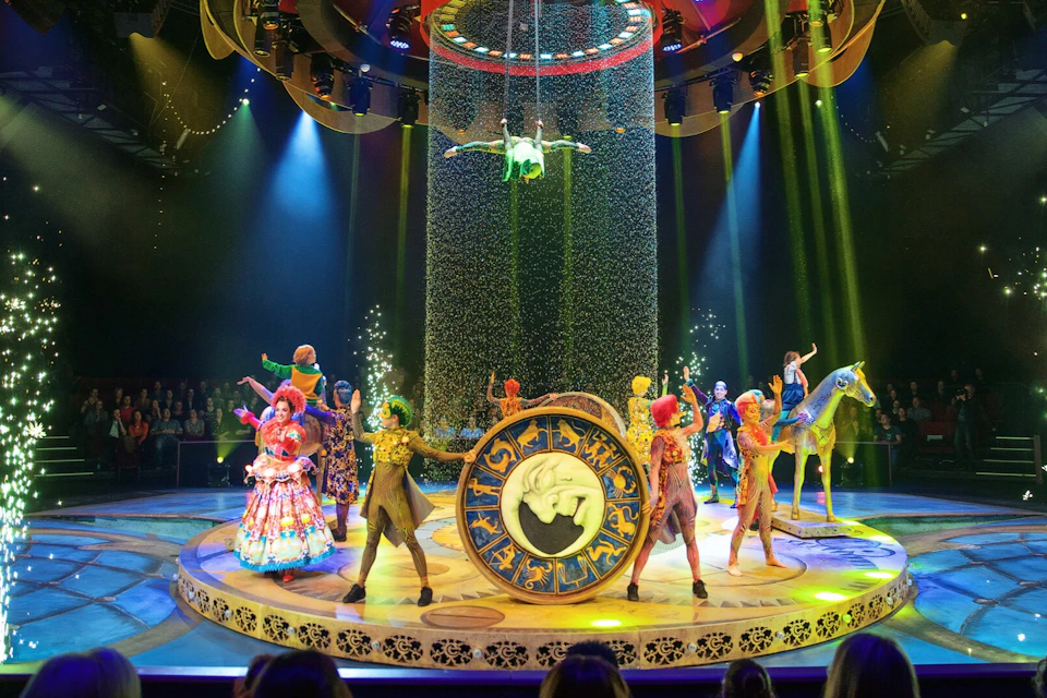 Performers in colorful costumes on a circular stage with an illuminated clock and an aerialist above, surrounded by an audience.