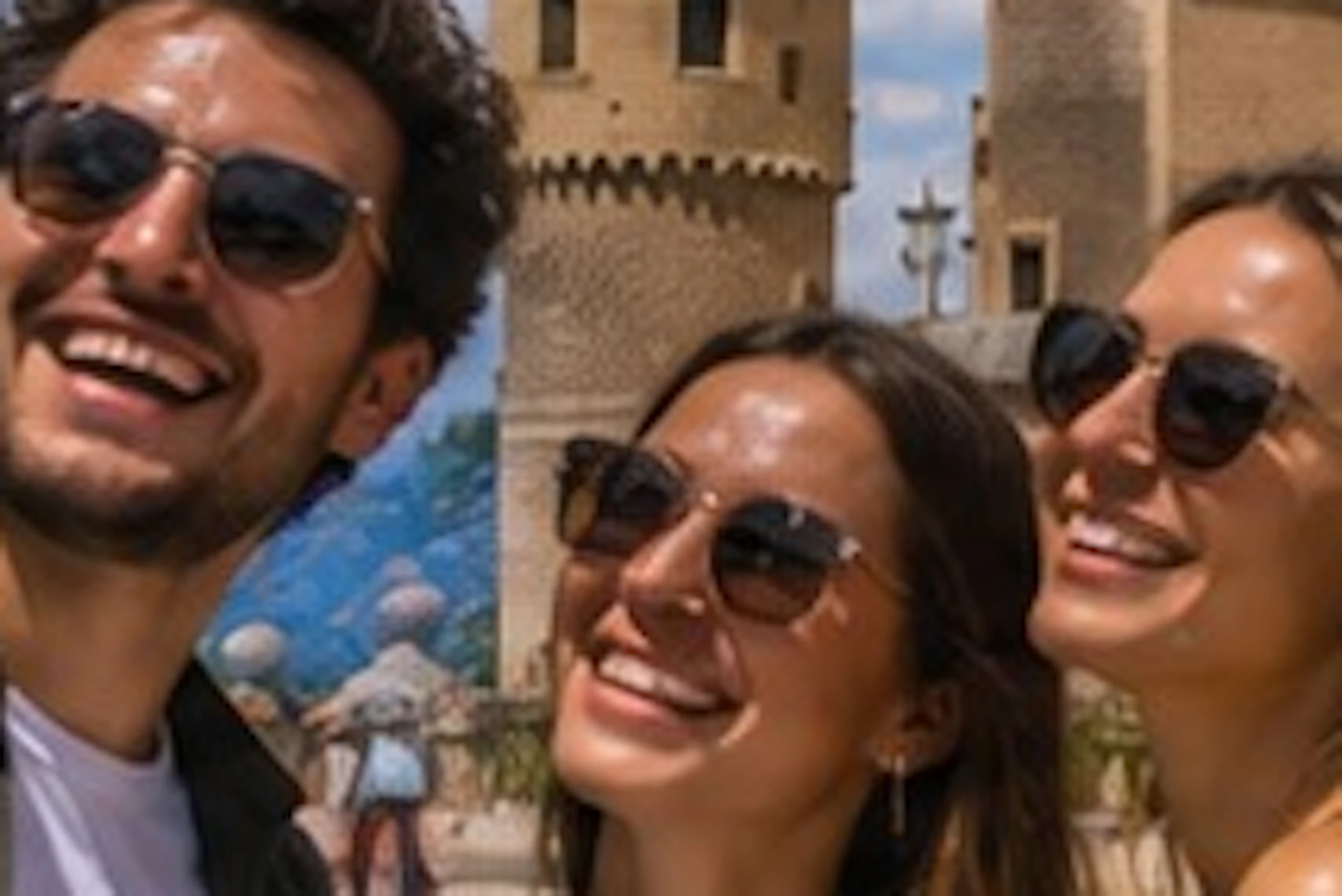 Three people smile for a selfie in front of a historic castle under a sunny, cloudy sky.