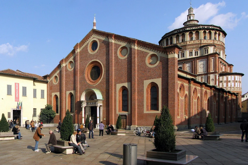 People sit and stand in front of a historic brick church with round windows and a dome on a sunny day.