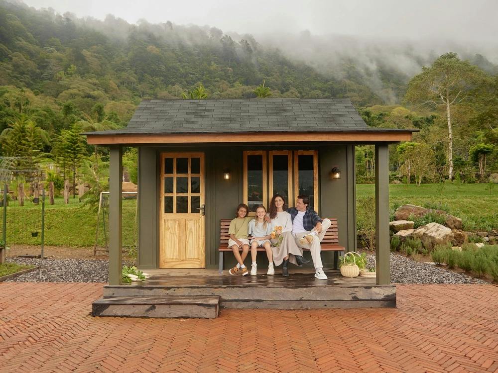A small house with a porch, where four people are seated together. The background shows lush greenery and misty hills.