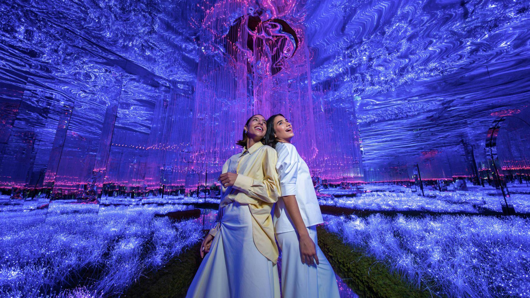 Twee vrouwen in lichtgekleurde outfits staan te midden van een verlichte, futuristische omgeving met gloeiende paarse en roze lichten.